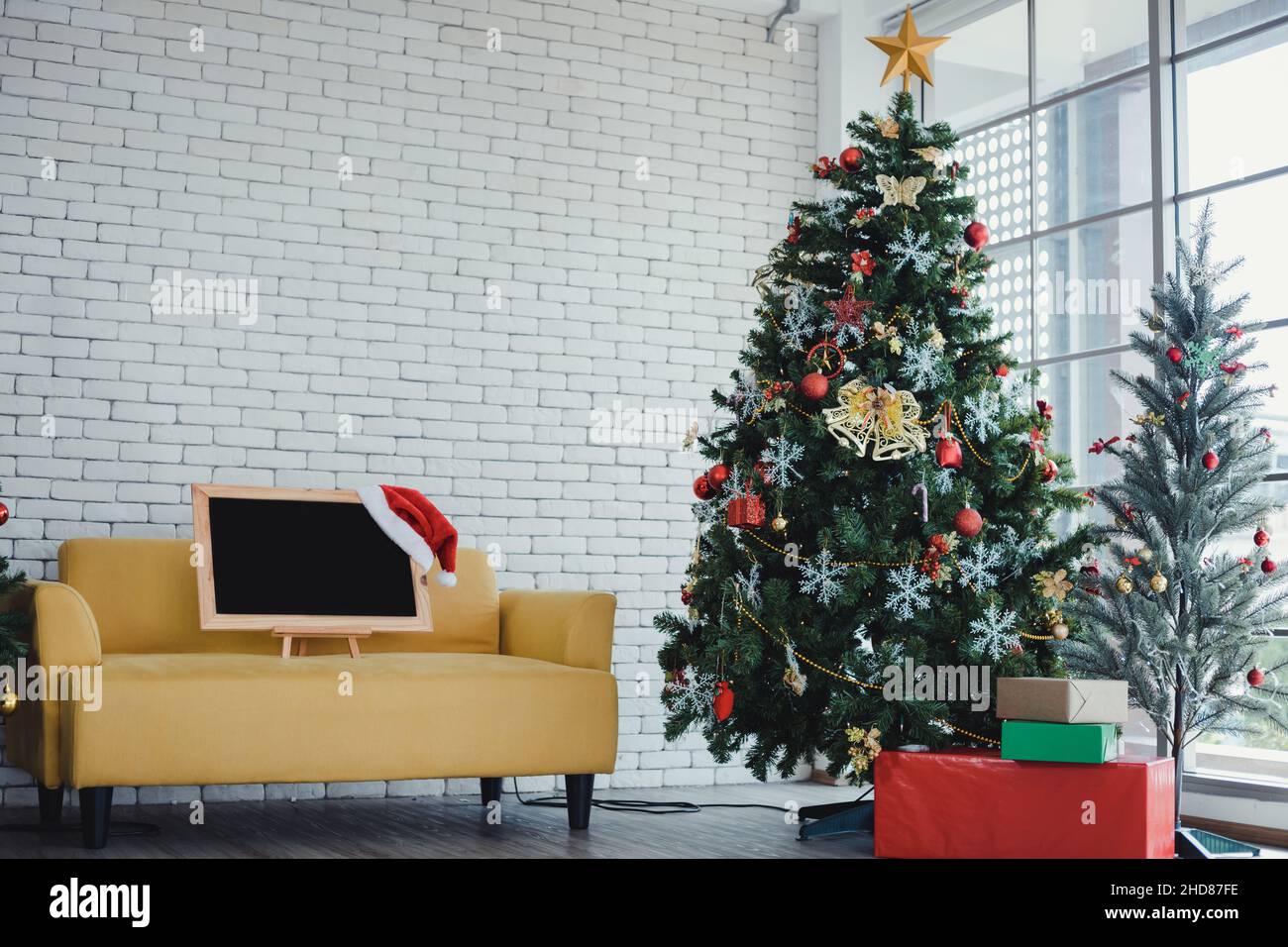 Tafel zum Einlegen von Text und einem roten Weihnachtsmann-Hut, auf gelbem Sofa mit weihnachtlichen Motiven im Raum zu Hause und Kopierraum dekoriert. Weihnachten und Stockfoto