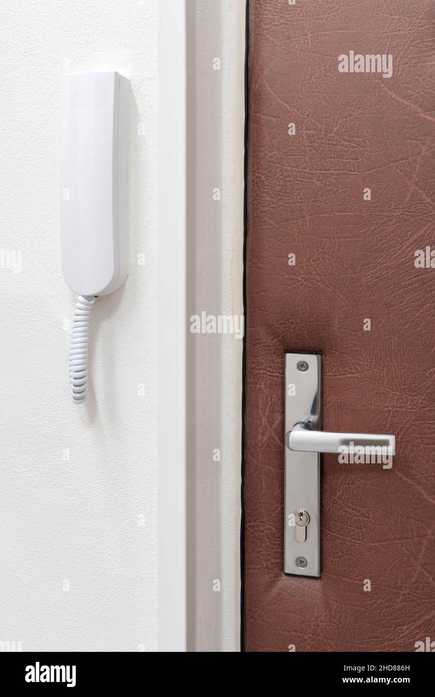Einfache weiße Gegensprechanlage oder Haustelefon an einer Wand neben einer geschlossenen Tür mit Metallgriff befestigt. Sicherheit und Kommunikation zu Hause. Stockfoto