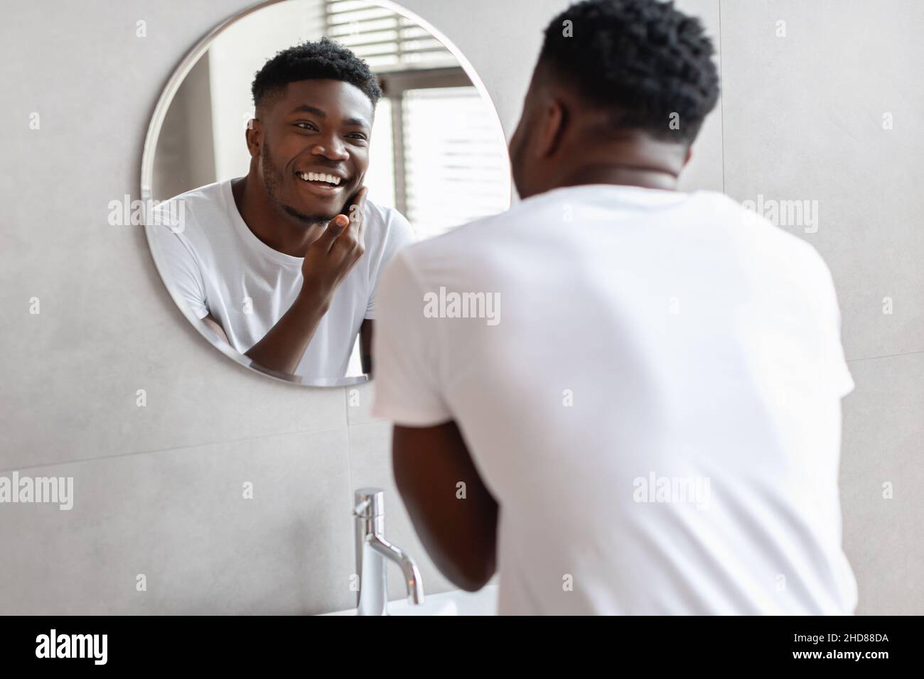 Schwarzer Mann Berührt Unrasiert Chin Lächelnd Auf Spiegel Im Badezimmer Stockfoto