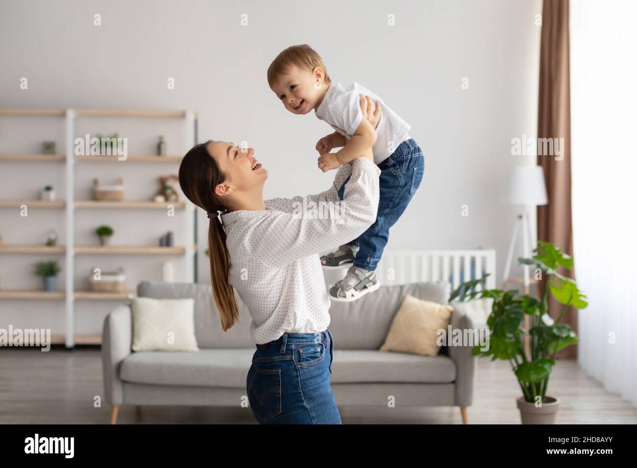 Lachende junge Mutter spielt mit niedlichen Kleinkind Sohn, hält lustige Kind vorgeblich fliegen, Spaß zusammen zu Hause Stockfoto