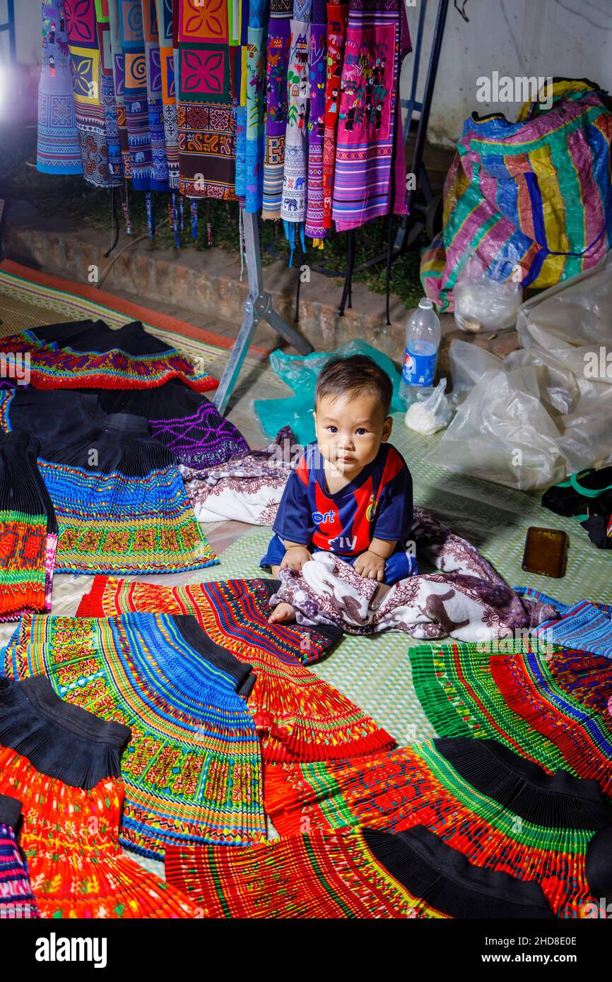 Ein süßer, einheimischer, molliger Junge sitzt in einem Verkaufsstand, der Stoffe auf dem Nachtmarkt in der Fußgängerzone im Zentrum von Luang Prabang, im Norden von Laos, Südostasien, verkauft Stockfoto