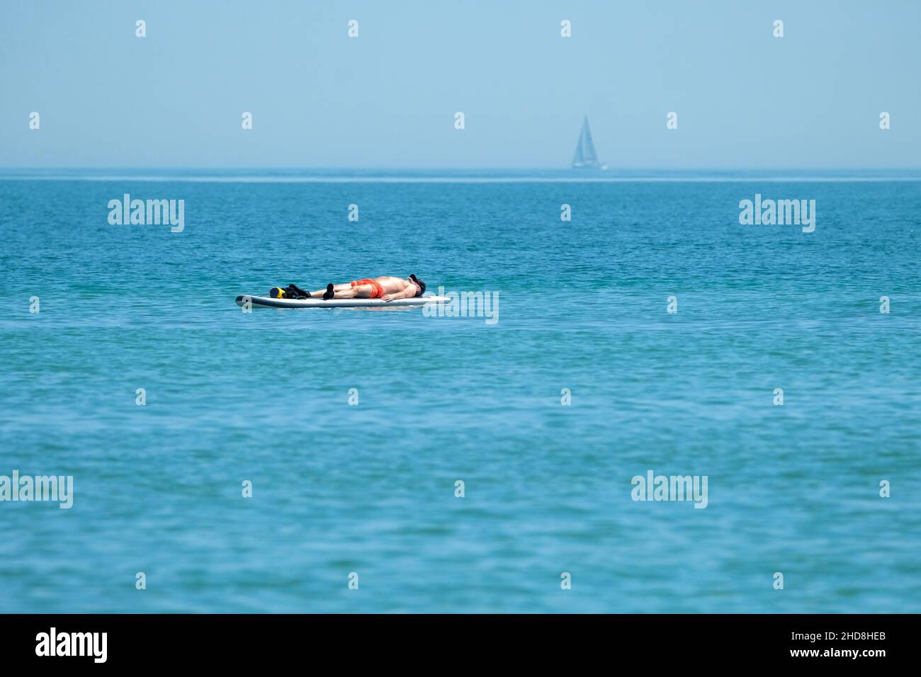 Mann, der auf einem Paddleboard auf einem ruhigen Meer mit einer Yacht im Hintergrund schwimmt, beim Sonnenbaden Stockfoto
