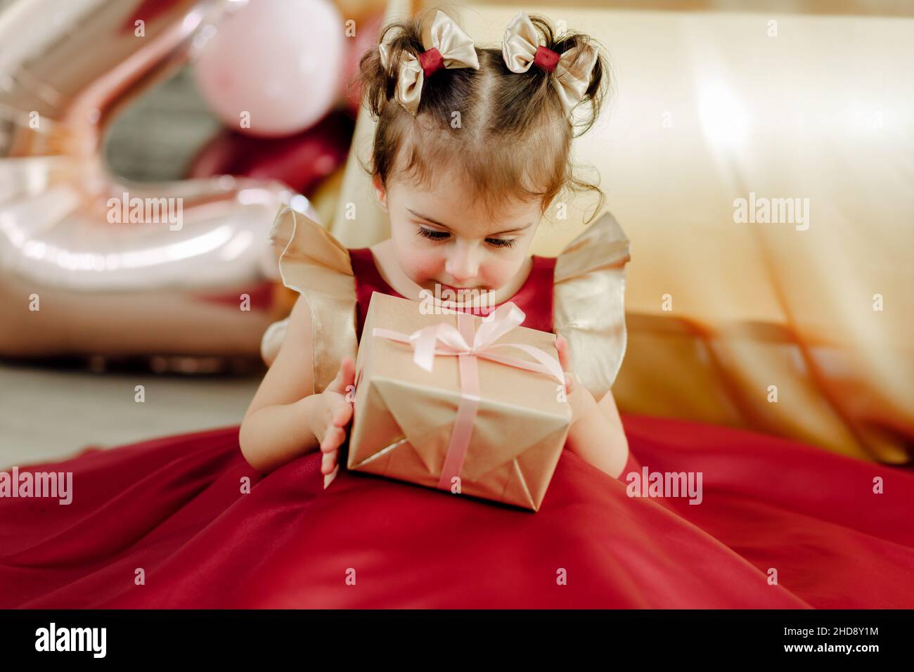 Staunend zwei Jahre altes Mädchen in einem roten Kleid hält und schaut sorgfältig auf ihr Geburtstagsgeschenk. Schöne Baby Mädchen Geburtstag Fotoshooting Stockfoto