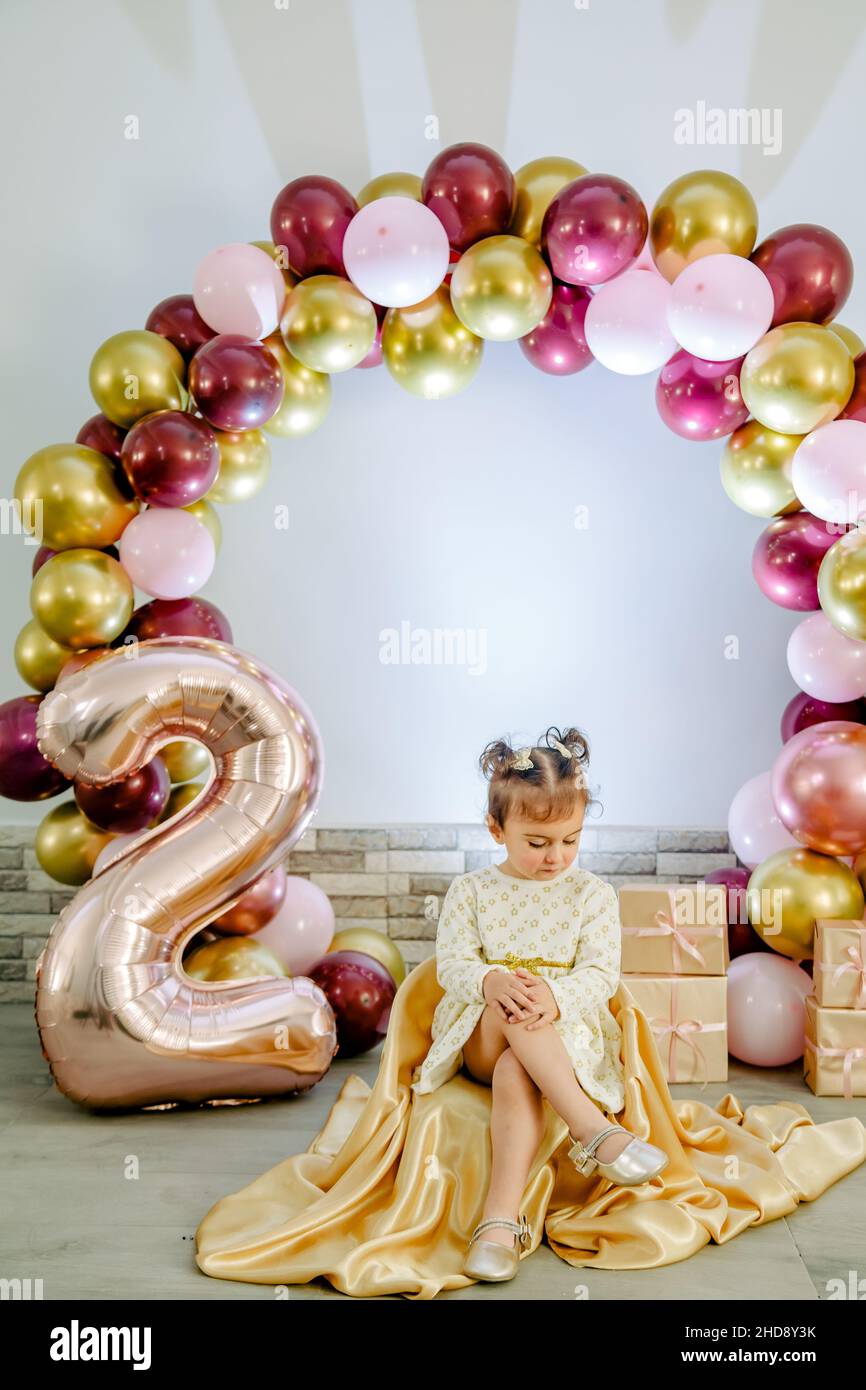 Modische Baby-Mädchen in einer weißen Kleidung feiert ihren zweiten Geburtstag posiert für die Kamera und schaut nach unten. Schöne Baby Mädchen Geburtstag Fotoshooting Stockfoto