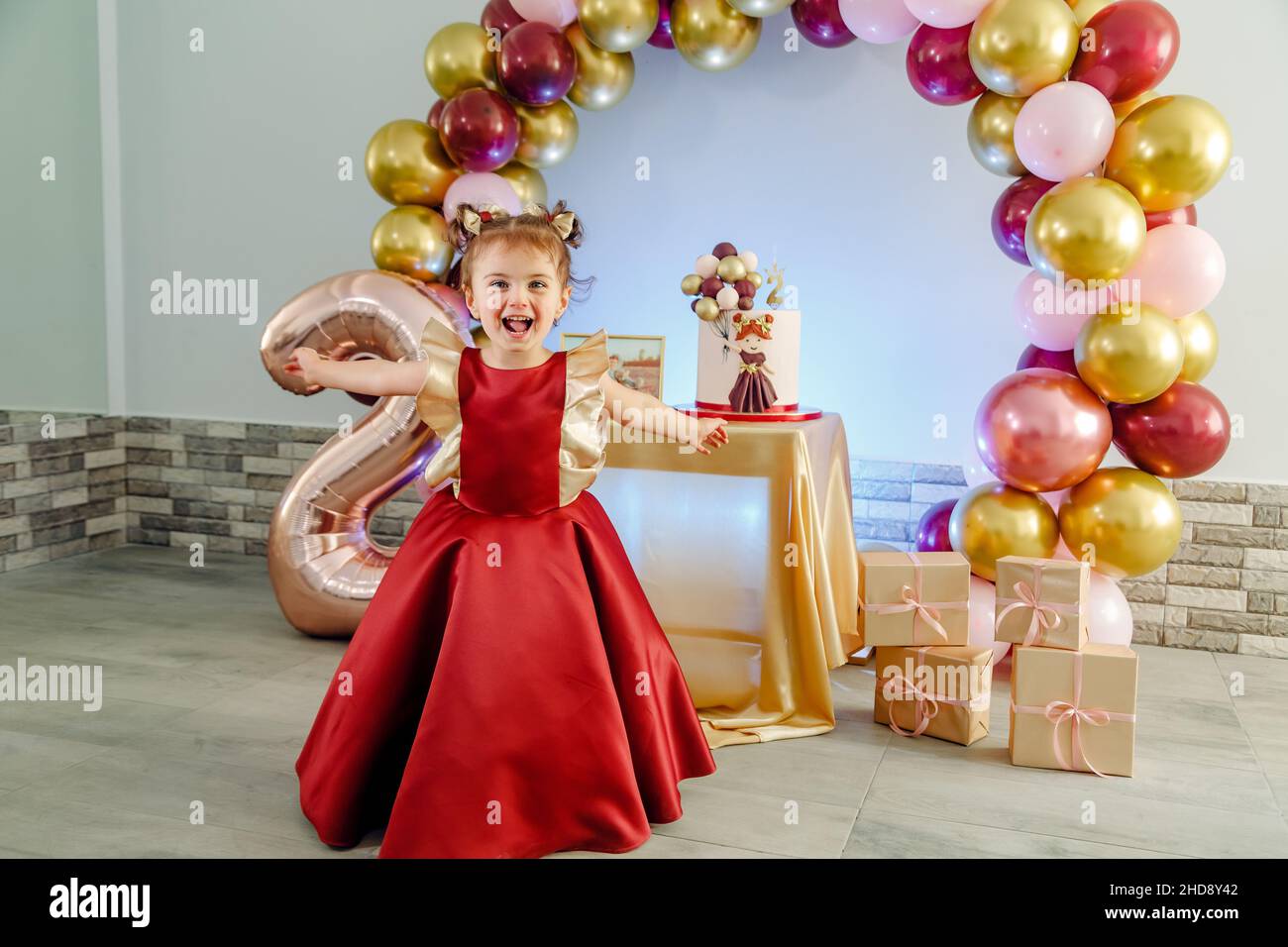 Lustige zwei Jahre alte Baby-Mädchen mit roten Kleidern Spaß vor ihrem Geburtstagskuchen. Schöne Baby Mädchen Geburtstag Fotoshooting Stockfoto