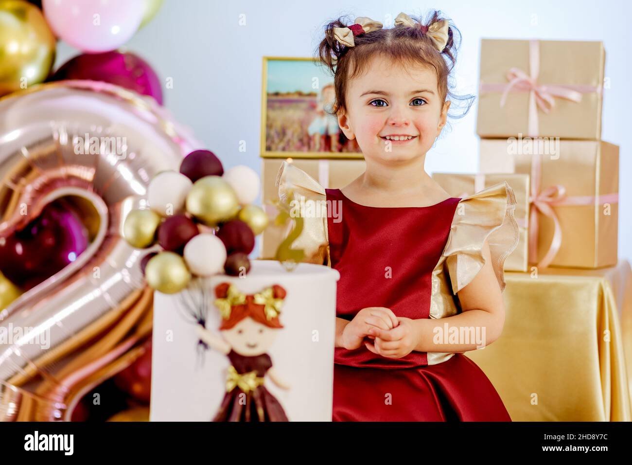 Zwei Jahre altes kleines lächelndes Mädchen, das neben dem Geburtstagskuchen steht und die Kamera aufmerksam anschaut. Schöne Baby Mädchen Geburtstag Fotoshooting Stockfoto