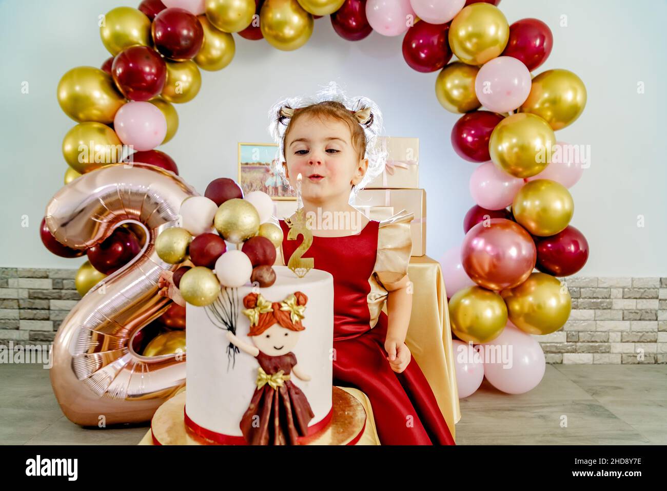 Zwei Jahre altes nettes und niedliches Mädchen in einem roten Kleid bläst Kerze auf einem Geburtstagskuchen aus. Schöne Baby Mädchen Geburtstag Fotoshooting Stockfoto