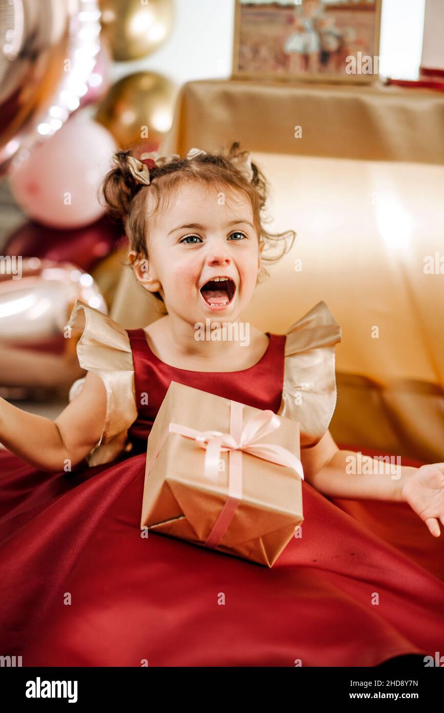 Vertikales Porträt von fröhlichen zwei Jahre alten Mädchen in einem roten Kleid mit ihrem Geburtstagsgeschenk. Schöne Baby Mädchen Geburtstag Fotoshooting Stockfoto