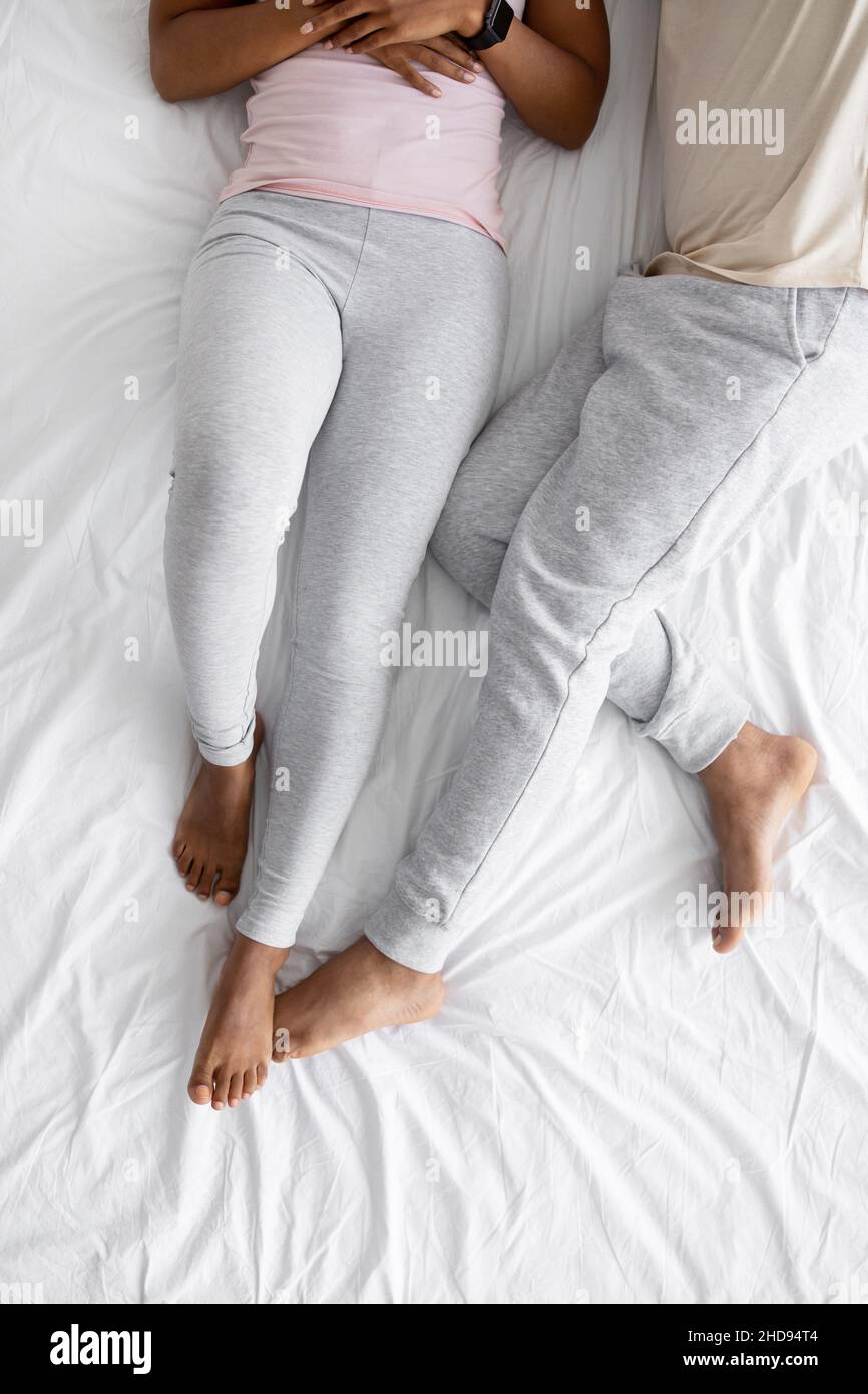 Das junge afroamerikanische Paar liegt auf weißem Bett in Hauskleidung, Draufsicht, nicht erkennbar, senkrecht Stockfoto