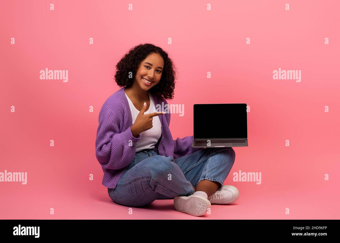 Positive junge schwarze Frau zeigt auf Laptop mit leerem Bildschirm, sitzend mit gekreuzten Beinen auf rosa Studio-Hintergrund, Nachbau Stockfoto