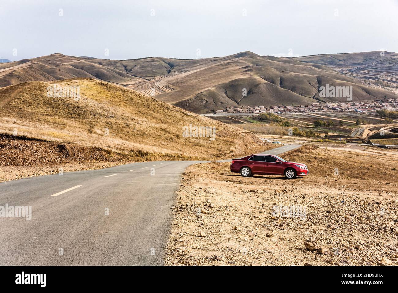 Das Auto steht an einer Straße zwischen goldenen Feldern, umgeben von Bergen unter einem bewölkten Himmel Stockfoto
