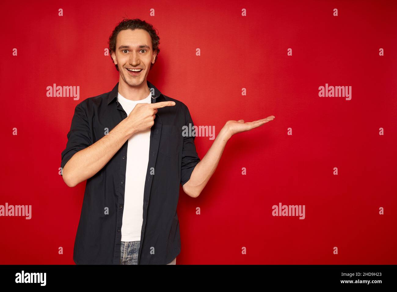 Junger, gutaussehender, aufgeregt Mann in Freizeitkleidung, blaues Hemd, weißes T-Shirt, zeigt Zeigefinger auf die Hand frei leeren Raum für Werbung Text Objekt isoliert roten Hintergrund Kopie Raum. Verkauf, promo. Hochwertige Fotos Stockfoto