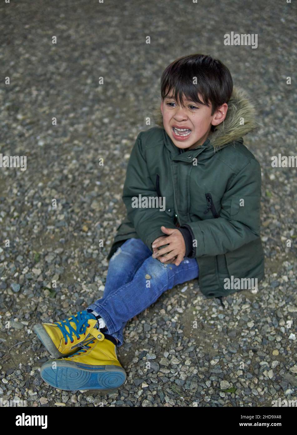 lateinisches Kind, das im Winter auf dem Boden sitzt und untröstlich mit offenem Mund im Freien weint, mit einem grünen Parka. Konzept der Frustration in der Kindheit Stockfoto