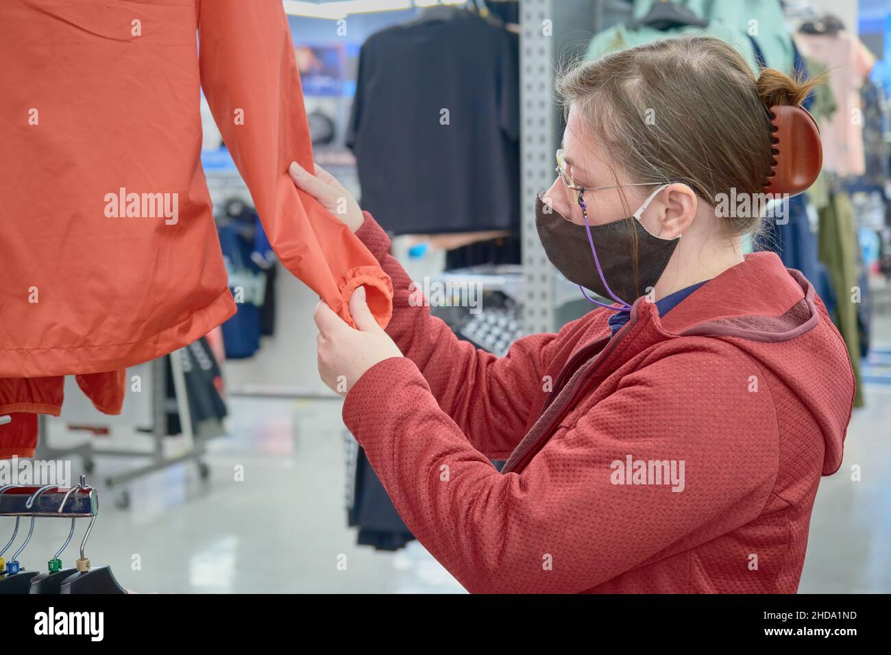 Kaukasische Frau in Maske gegen Virus wählt einen roten Windbreaker, um im Geschäft zu kaufen Stockfoto