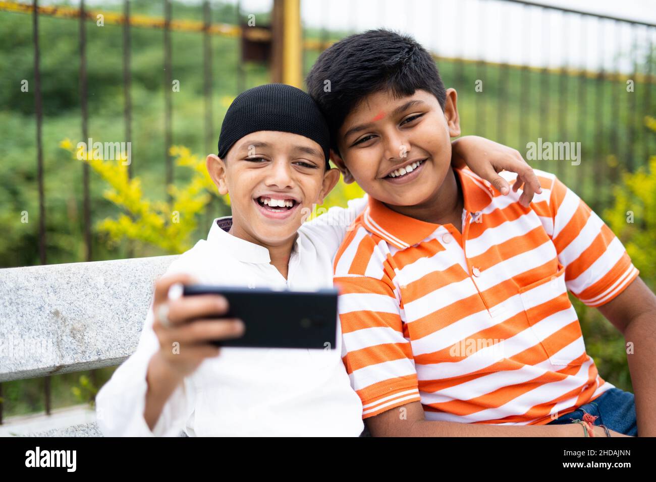 Zwei glückliche, multiethnische Kinder, die im Park Selfie auf dem Handy machen – ein Gepräch aus Freundschaft, religiöser Vielfalt und sozialer Harmonie Stockfoto