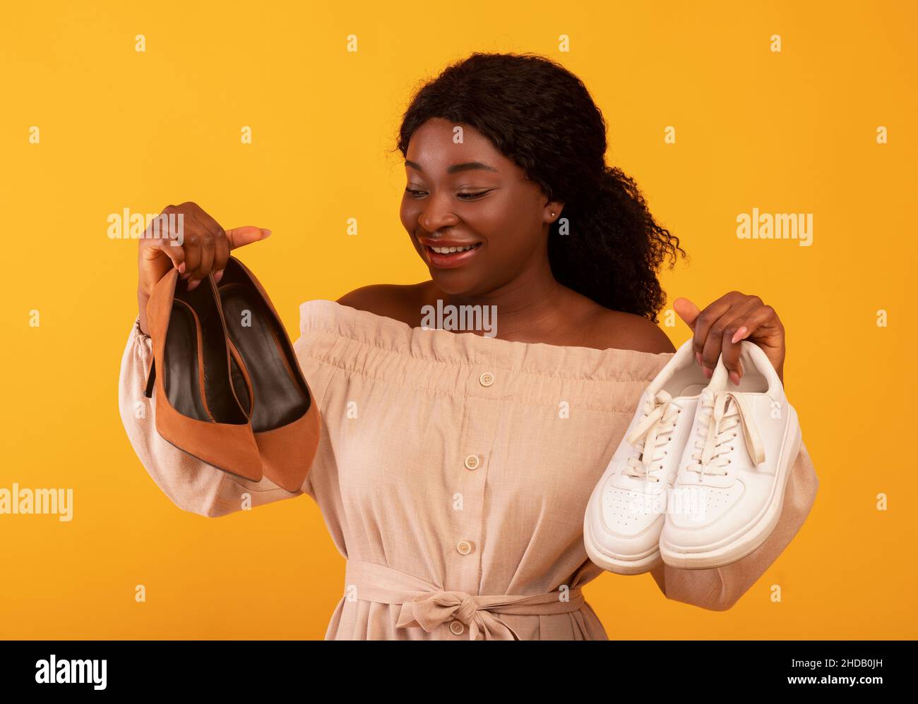 Junge afroamerikanische Frau, die zwischen Schuhen mit hohem Absatz und bequemen Sneakern auf orangefarbenem Hintergrund wählt Stockfoto