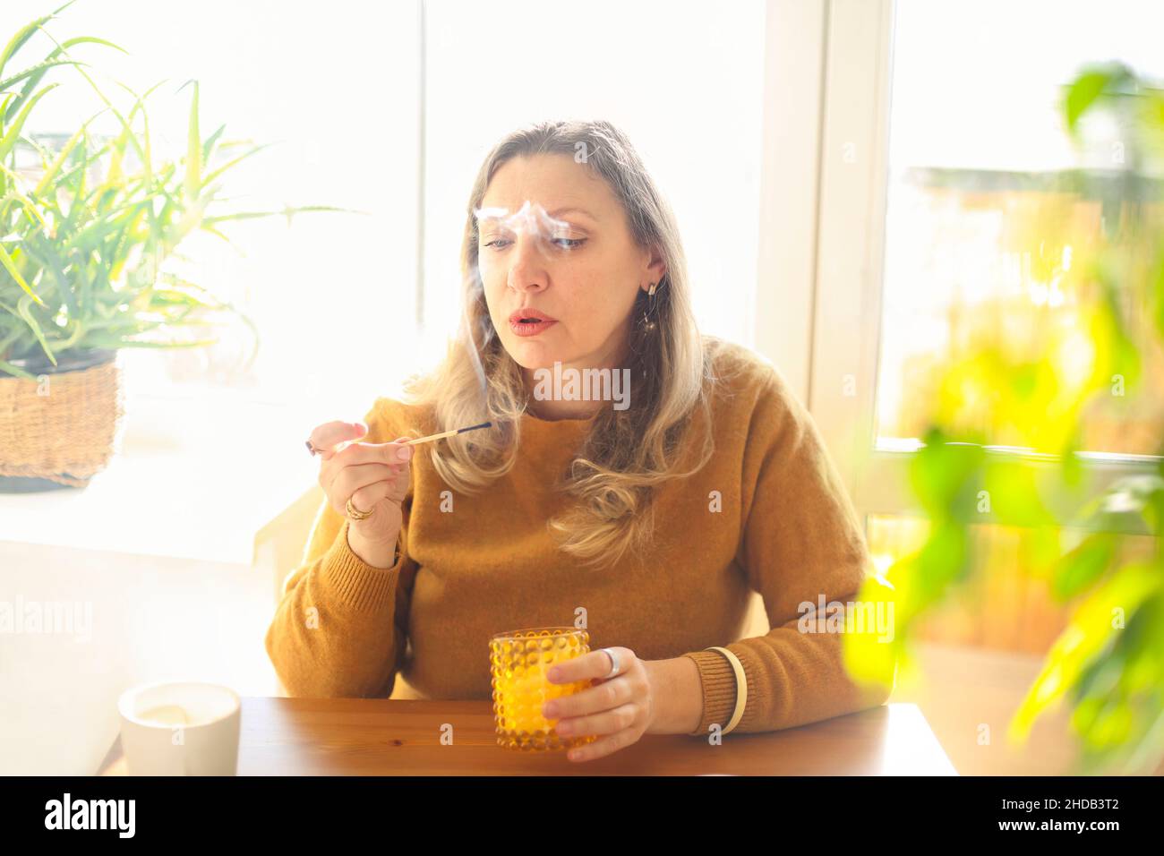 Friedliche, ruhige Frau im mittleren Alter von 40s Jahren mit brennender Duftkerze, während sie am Tisch in einem gemütlichen Zimmer zu Hause sitzt und eine warme, intime Atmosphäre schafft Stockfoto