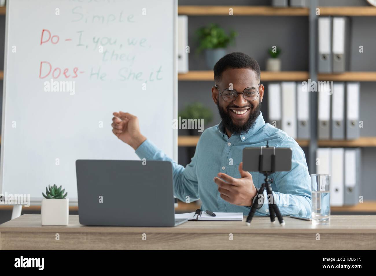 Lächelnd hübscher junger schwarzer männlicher Tutor mit Bart zeigen englische Regeln und schaut auf pc und Smartphone Stockfoto