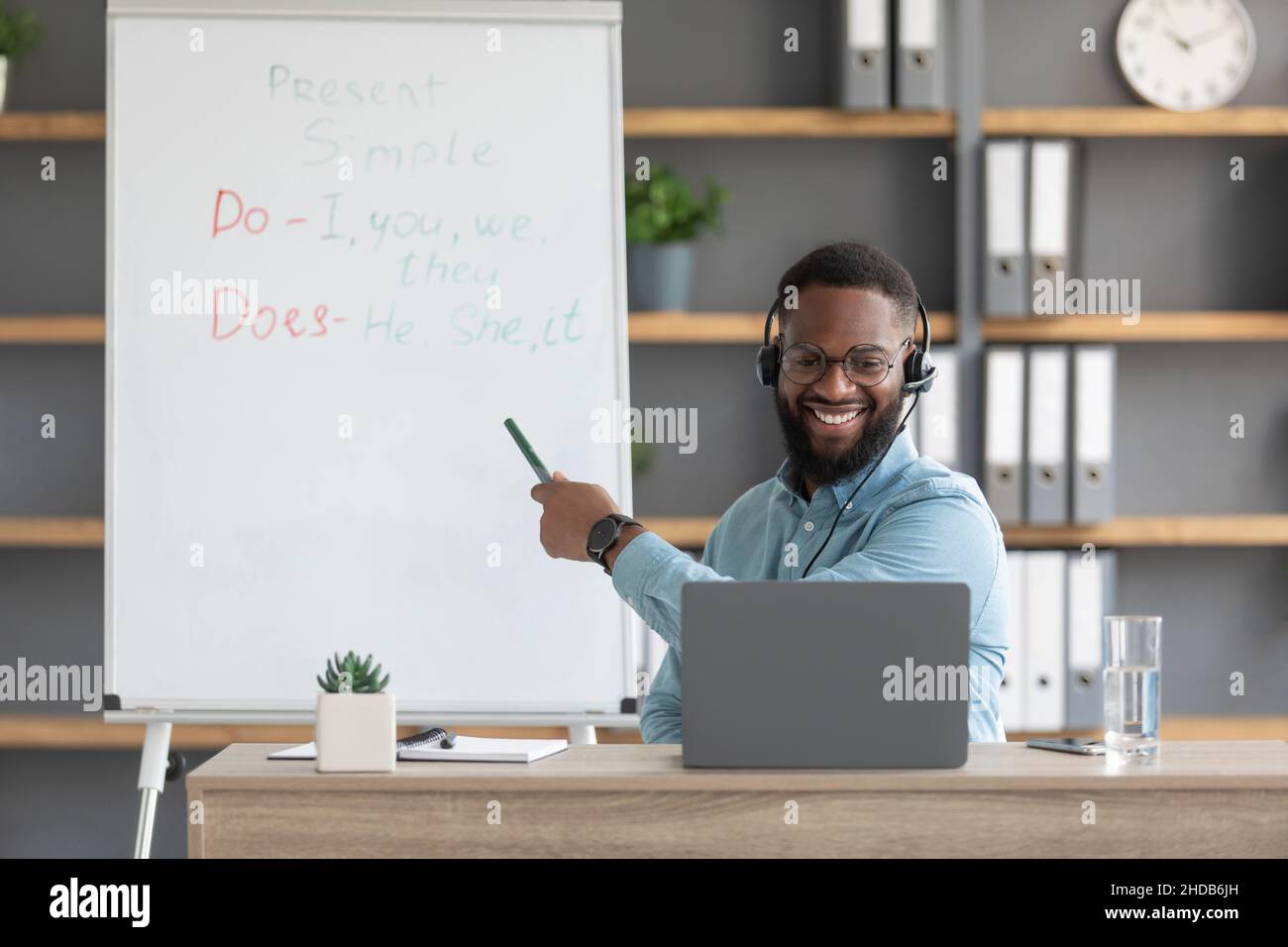Freundlicher junger afroamerikanischer Mann Tutor zeigt Regeln des englischen an der Tafel und schaut auf den pc Stockfoto