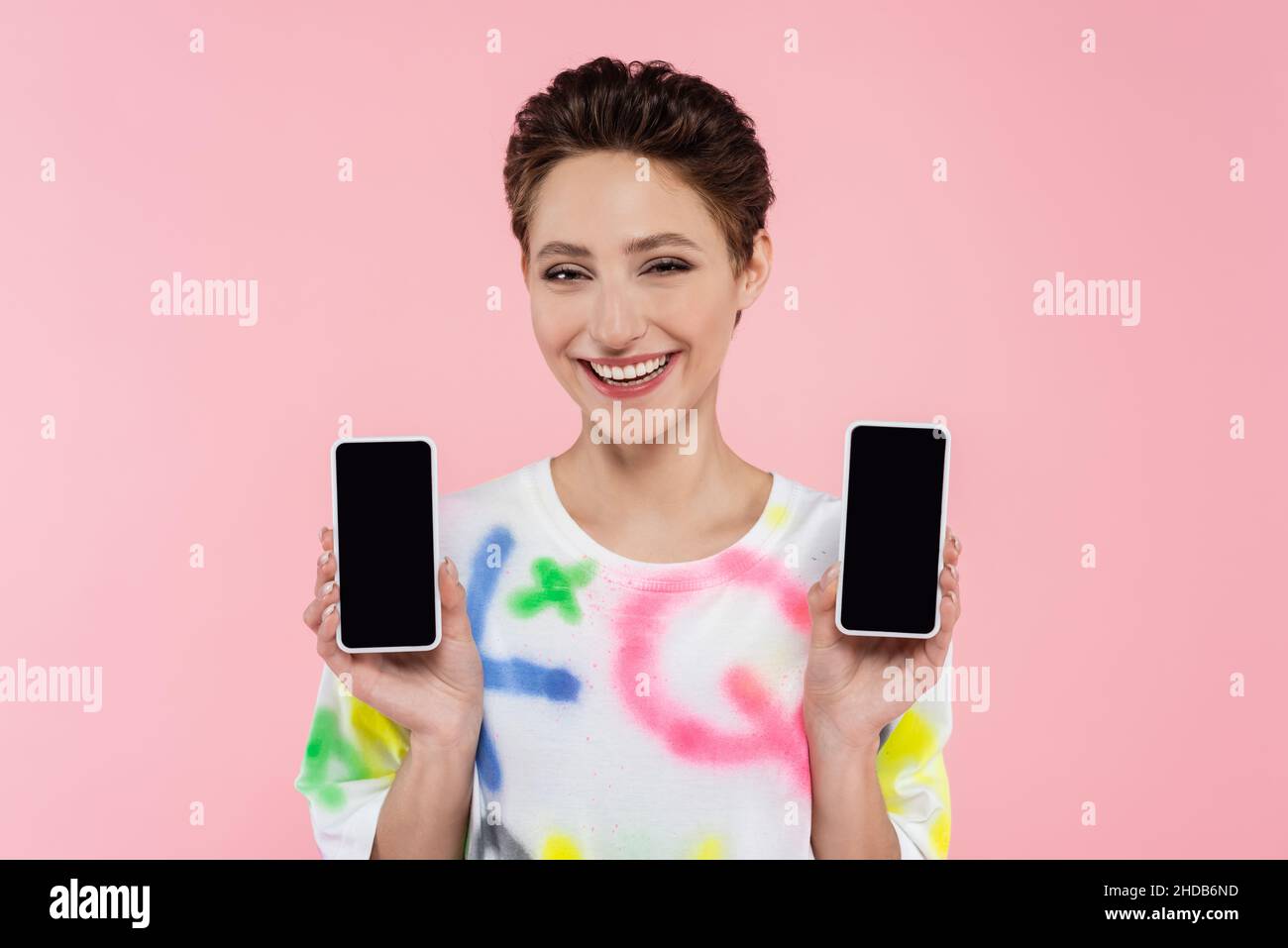 Junge Brünette Frau zeigt Handys, während lächelnd auf Kamera isoliert auf rosa Stockfoto