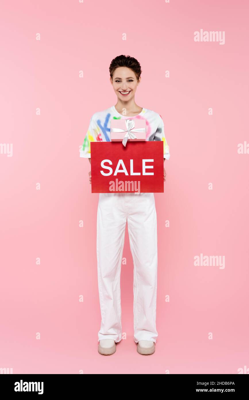 Durchgehende Ansicht der glücklichen Frau in weißer Hose mit Geschenkbox und roter Karte mit Verkaufsaufdruck auf rosa Stockfoto