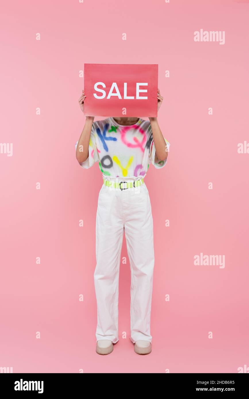 Lange Ansicht der Frau in weißen Hosen versteckt Gesicht hinter Karte mit Verkauf Schriftzug auf rosa Stockfoto