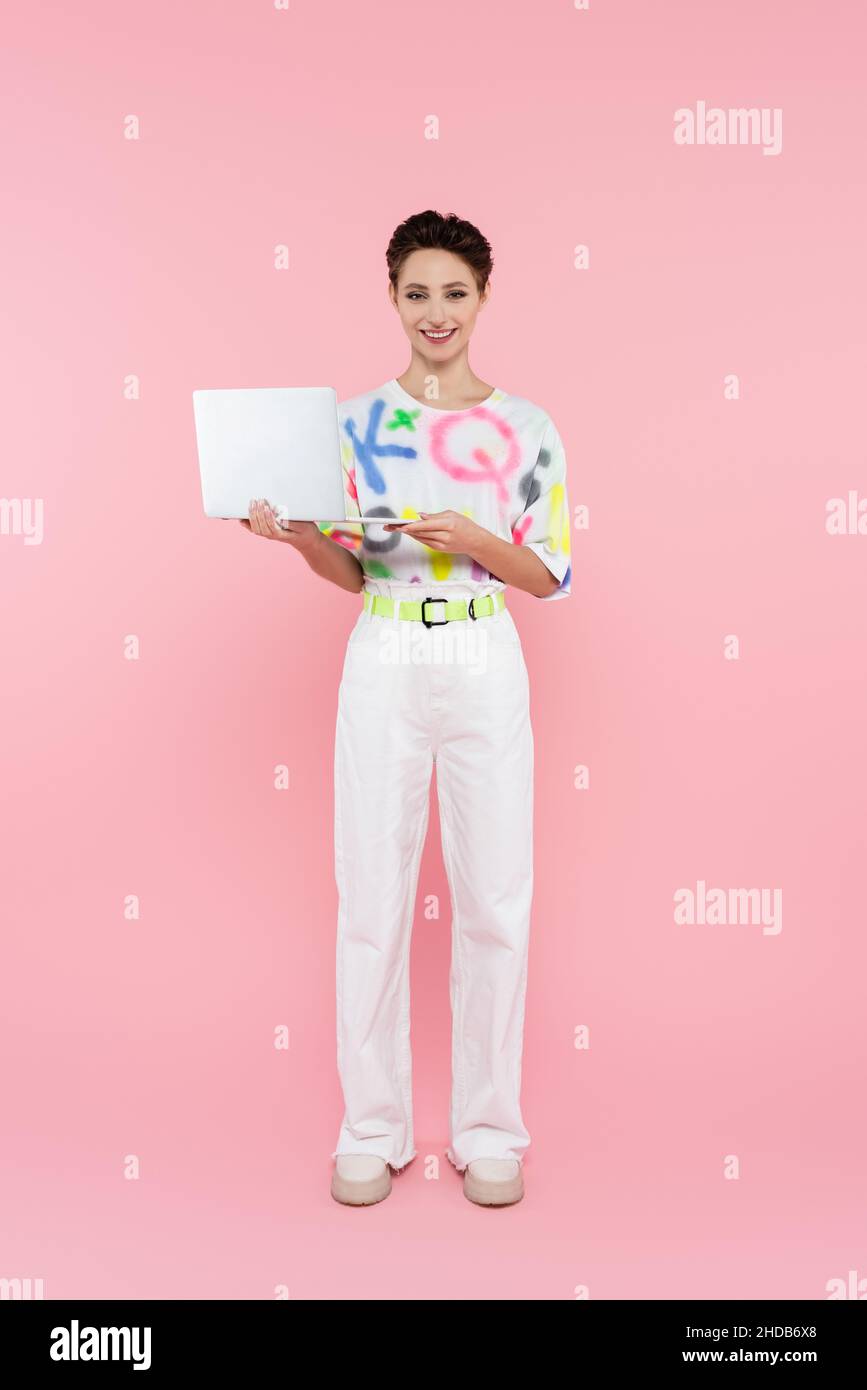 Lange Ansicht der erfreut Frau in weißen Hosen stehend mit Laptop auf rosa Stockfoto