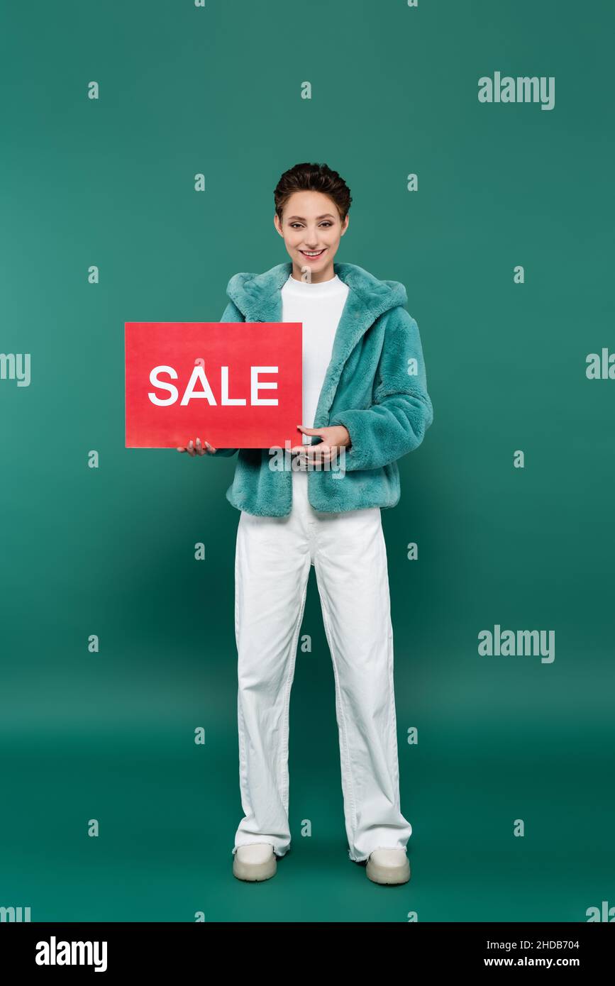 Lange Ansicht der glücklichen Frau in weißen Hosen und Kunstpelzjacke mit roter Karte mit Sale-Schriftzug auf grün Stockfoto