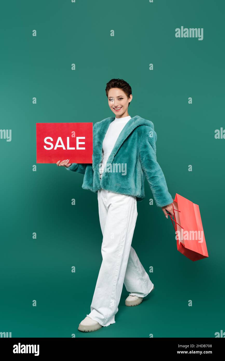 Fröhliche Frau in weißen Hosen und Kunstpelzjacke zu Fuß mit Verkaufskarte und Einkaufstasche auf grün Stockfoto