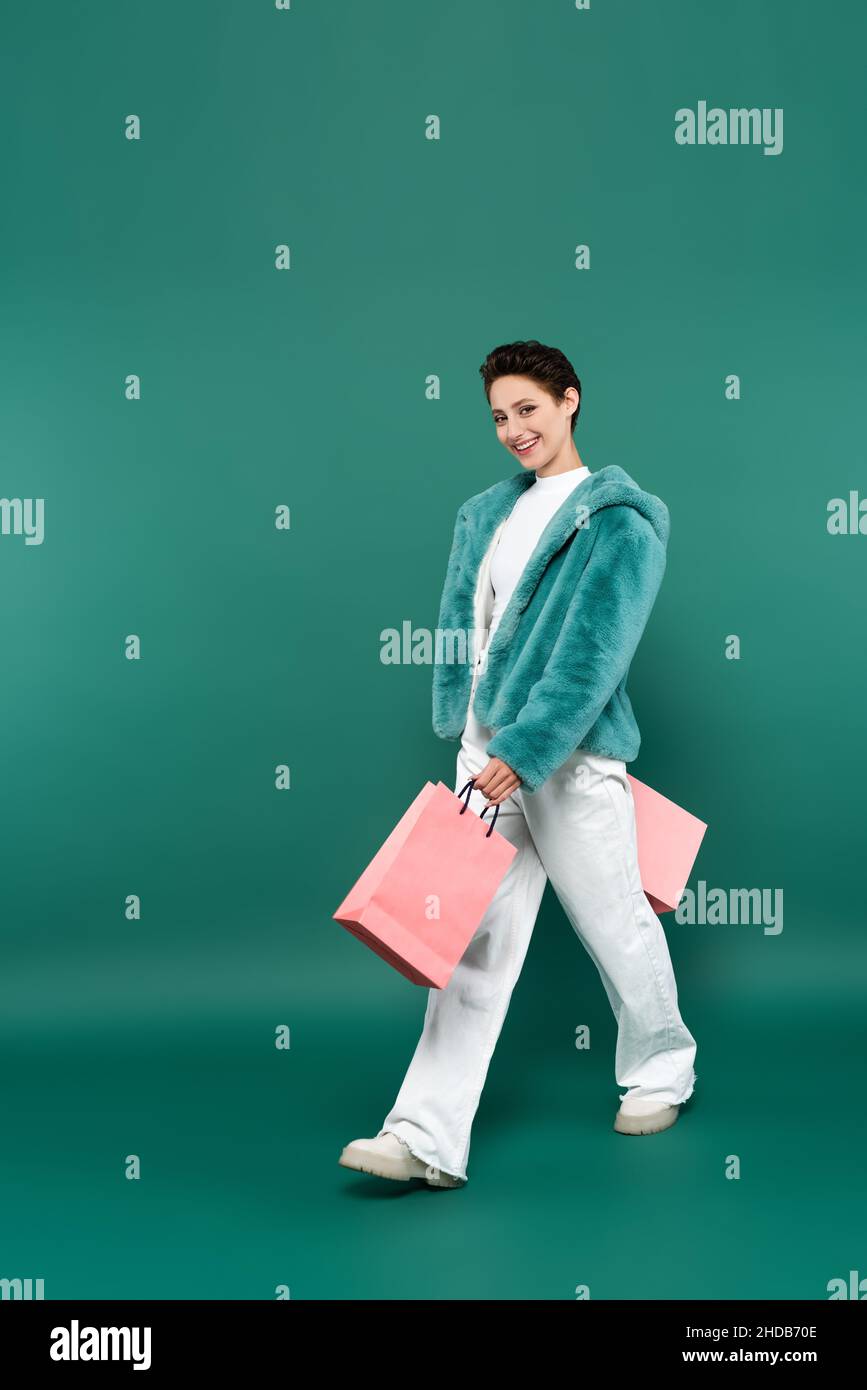 Fröhliche Frau in Kunstpelzjacke und weißer Hose, die mit pinken Einkaufstaschen auf Grün läuft Stockfoto