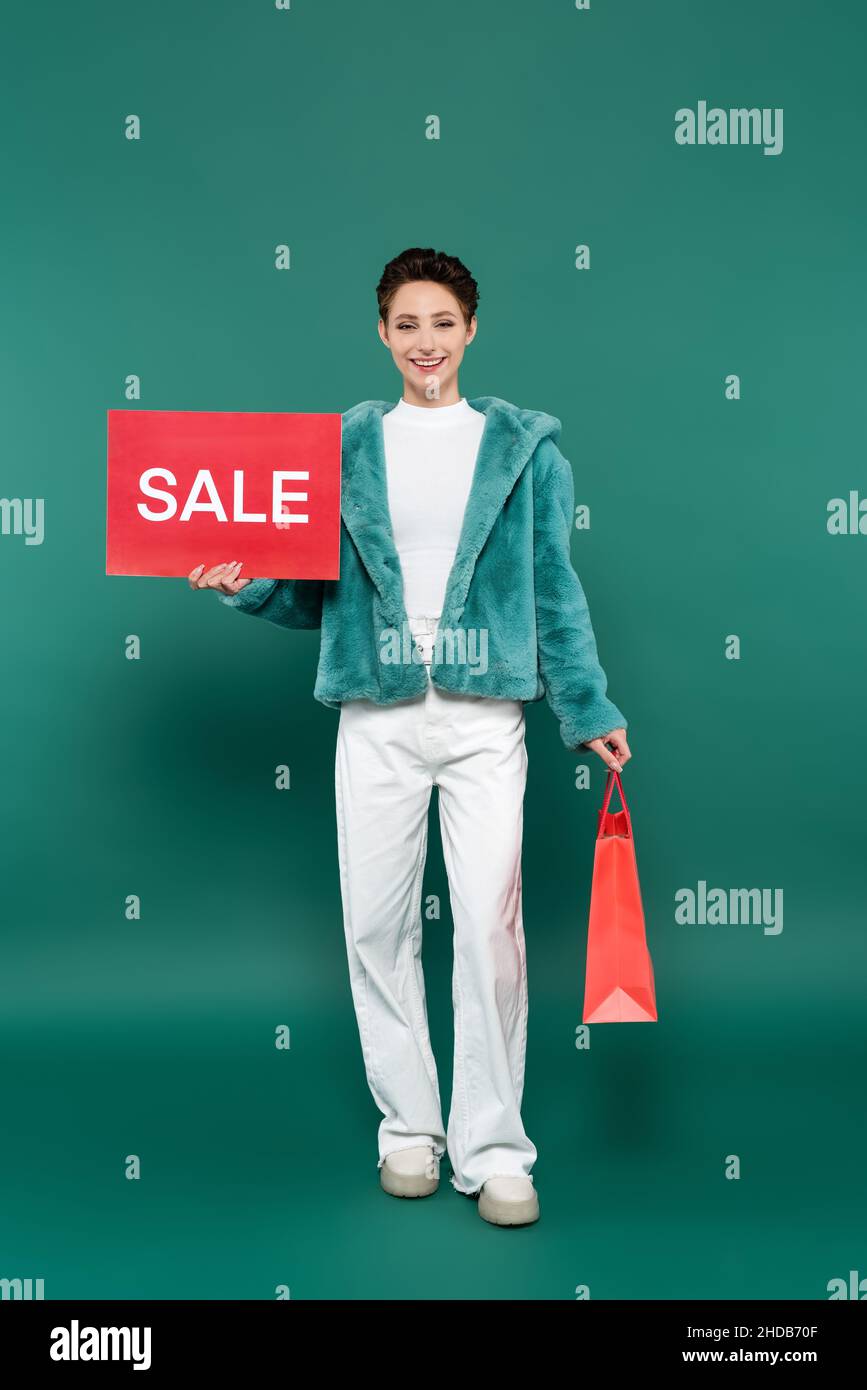 Fröhliche Frau in Kunstpelzjacke und weißer Hose mit Einkaufstasche und roter Karte mit Sale-Schriftzug auf Grün Stockfoto