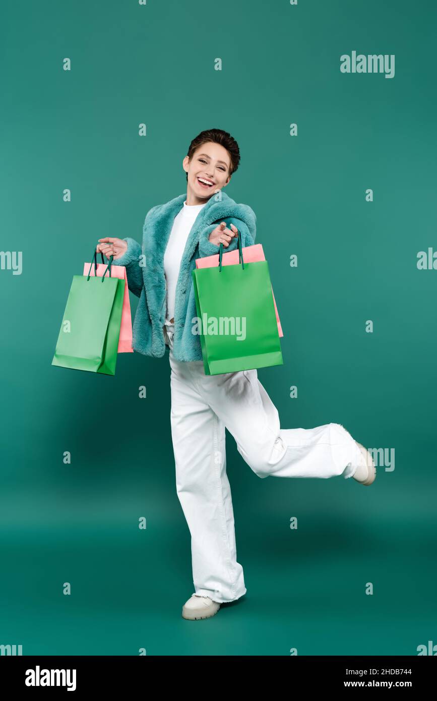 Lange Ansicht der aufgeregten Frau in Kunstpelzjacke und weißen Hosen posiert mit Einkaufstaschen auf grün Stockfoto