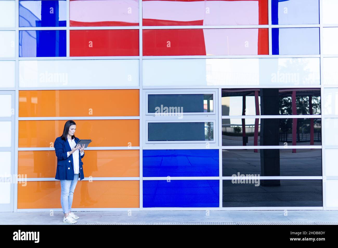 Schöne kaukasische Geschäftsfrau mit einem Tablet vor einem Bürogebäude. Sie trägt eine lässige blaue Jacke. Farbenfroher Hintergrund Stockfoto