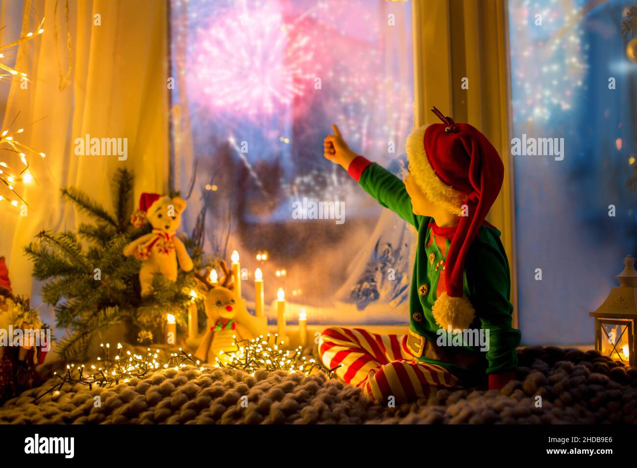 Niedliches Kleinkind in Pyjama, das auf einer wütsamen Decke neben einem Fenster sitzt und draußen auf ein Feuerwerk am Silvesterabend blickt Stockfoto