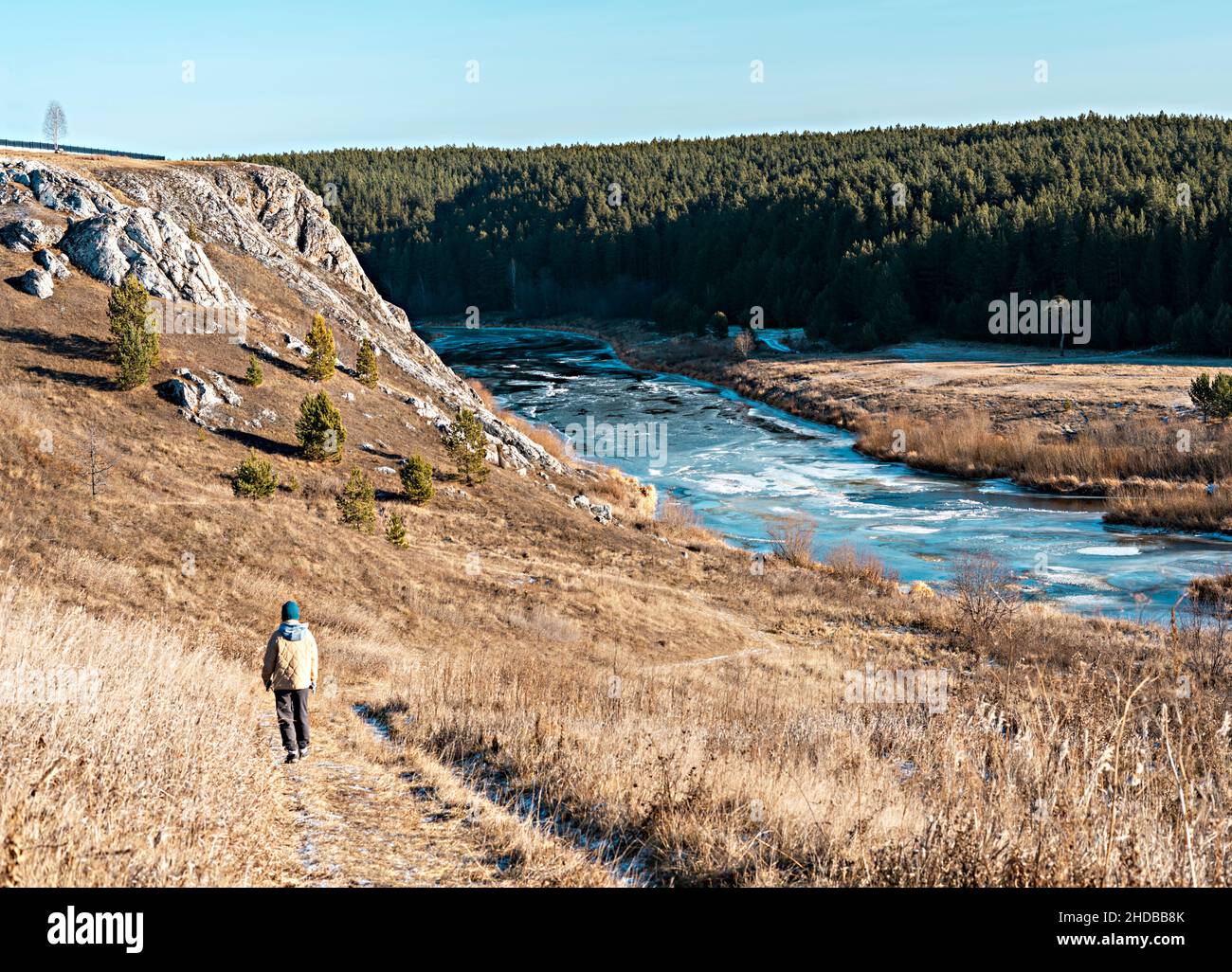 Rückansicht einer Frau in neutralen Farben Kleidung, die im Herbst auf trockenem Gras gegen Felsen und Wald am Fluss entlang läuft, Landschaft in Erdtönen, Wandern genießen Stockfoto