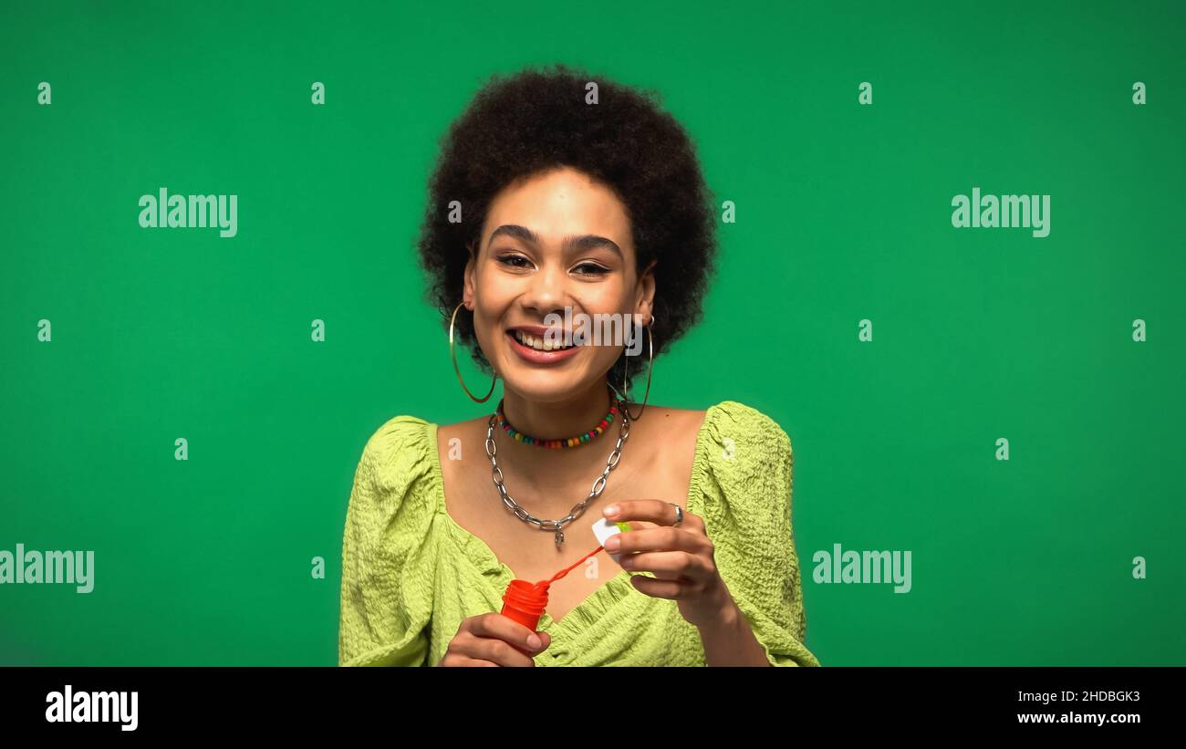 Glückliche afroamerikanische Frau, die eine Flasche mit Seifenblasen und Seifenblasen auf grün isoliert hält Stockfoto