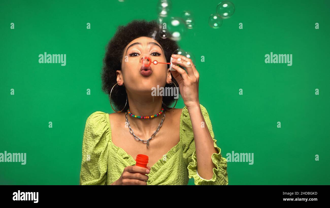 Lockige afroamerikanische Frau bläst Seifenblasen, während sie den Seifenstab isoliert auf Grün hält Stockfoto
