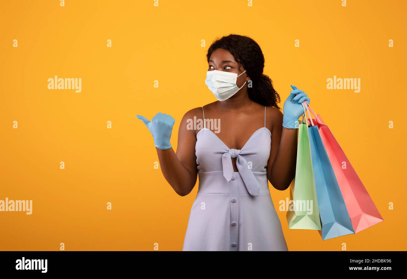 Tausendjährige schwarze Frau in Anti-Covid-Maske und Gummihandschuhen mit Einkaufstaschen, die auf Leerraum zeigen Stockfoto