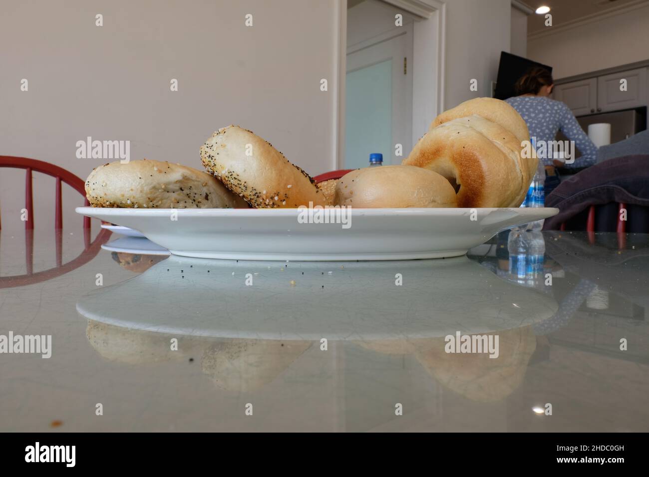 Seitenansicht eines weißen Keramiktellers auf einer Glastafel mit verschiedenen Bagels; authentischer Küchentisch mit Frühstücksküche. Stockfoto