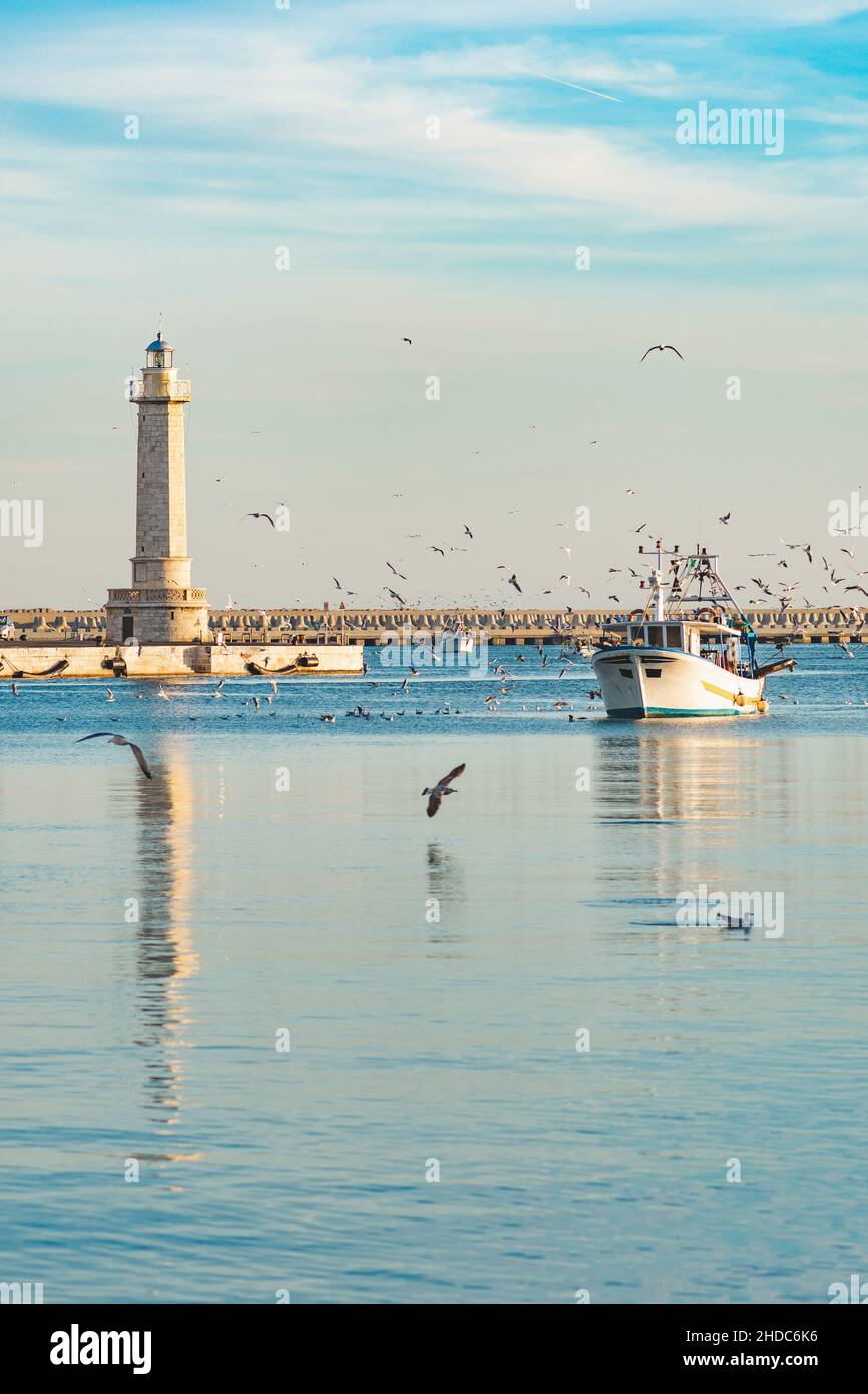 Altes Fischerboot, das vom Fischfang zurückkehrt, kommt bei Sonnenuntergang in den Hafen, begleitet von einer Schar von Möwen. Mit schönem Leuchtturm im Hintergrund Stockfoto