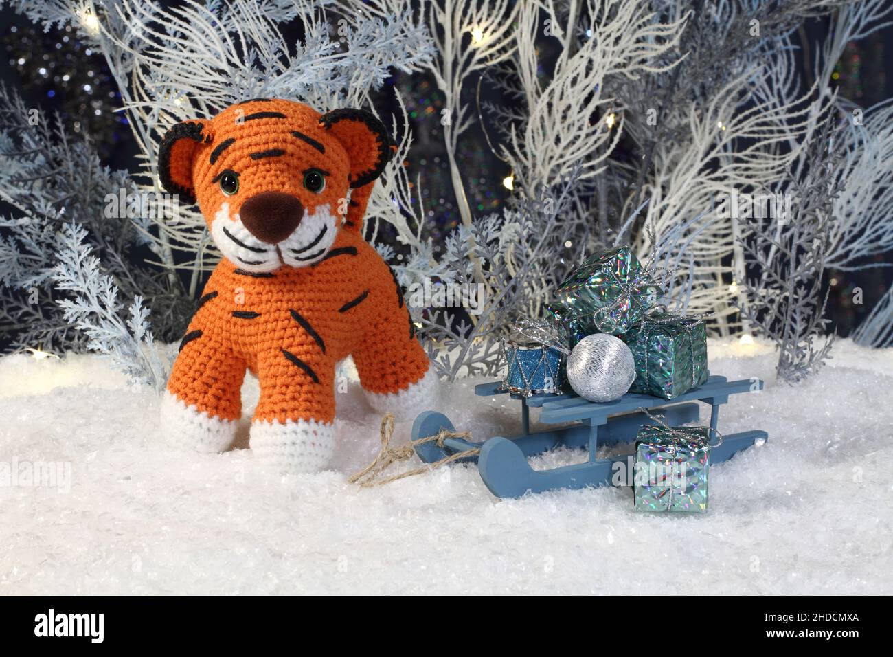 Ein gestrickter Spielzeugtiger steht auf dem künstlichen Schnee und ein Spielzeugschlitten mit Geschenken steht neben ihm vor dem Hintergrund weißer und blauer Kunstbäume Stockfoto