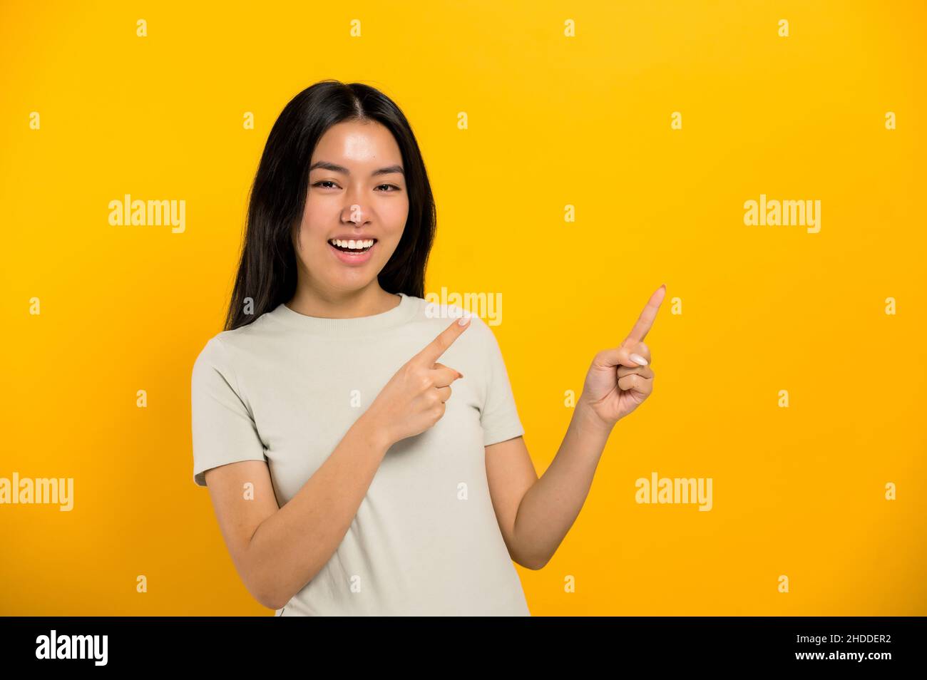 Happy lovely Millennial Brunette langhaarige asiatische junge Frau, zeigt mit den Händen auf leeren Raum, blickt auf die Kamera, lächelnd freundlich, steht auf isoliertem Hintergrund in lässigem T-Shirt Stockfoto