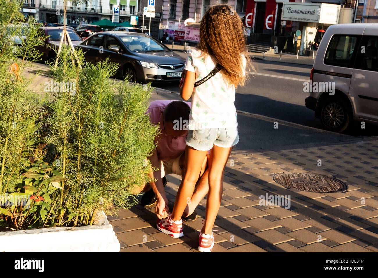 Papa hilft der Tochter, an sonnigen Sommertagen auf einer Straße in Kiew Schnürsenkel an ihren Sneakers zu binden. Mädchen nimmt Hilfe mit wunderbarer Stille und königlicher Anmut an. Stockfoto
