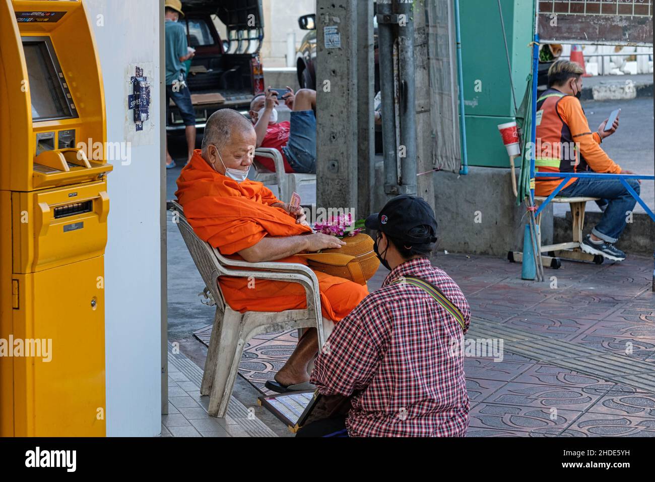 Ein buddhistischer Mönch mit orangenen Roben, der neben einem gelben Geldautomaten sitzt, überlegt, welche Lottoscheine von einem Lotteriehändler gekauft werden sollten; Bangkok, Thailand Stockfoto