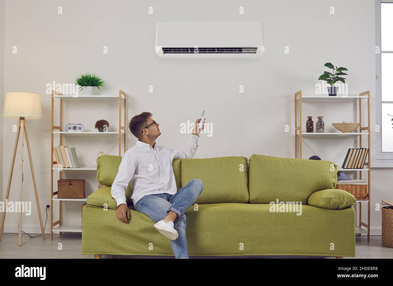 Der Mann, der zu Hause auf dem Sofa sitzt, schaltet per Fernbedienung die Klimaanlage an der Wand ein Stockfoto