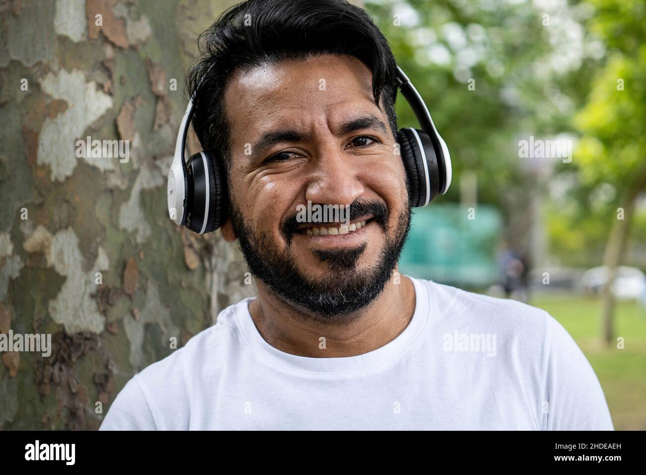 Portrait lateinischer bärtiger Mann mit Kopfhörern, der die Kamera anlächelt und im Park Musik hört Stockfoto