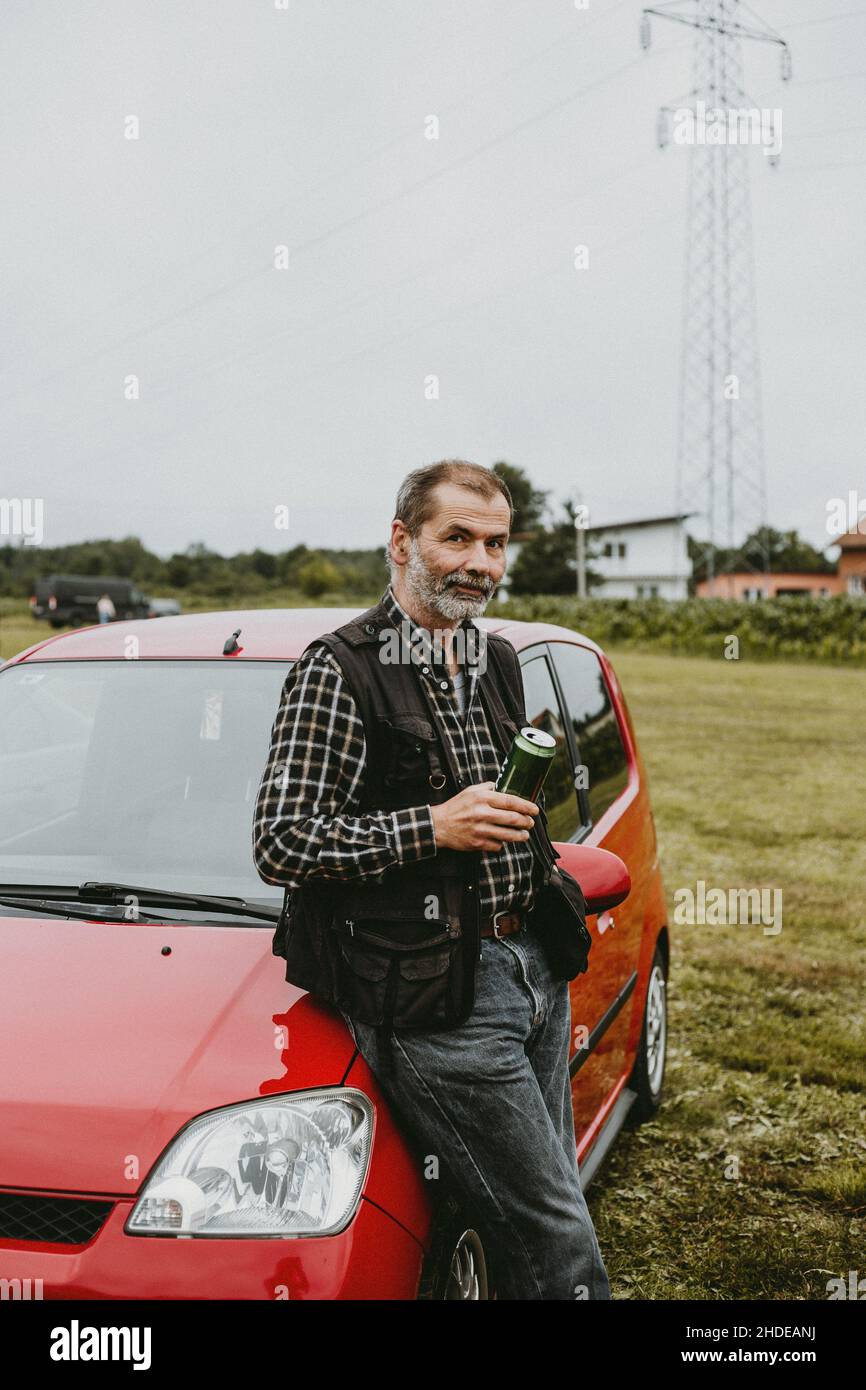 Älterer Mann, der vor dem Auto steht und eine Dose eisgekühltes Bier hält Stockfoto