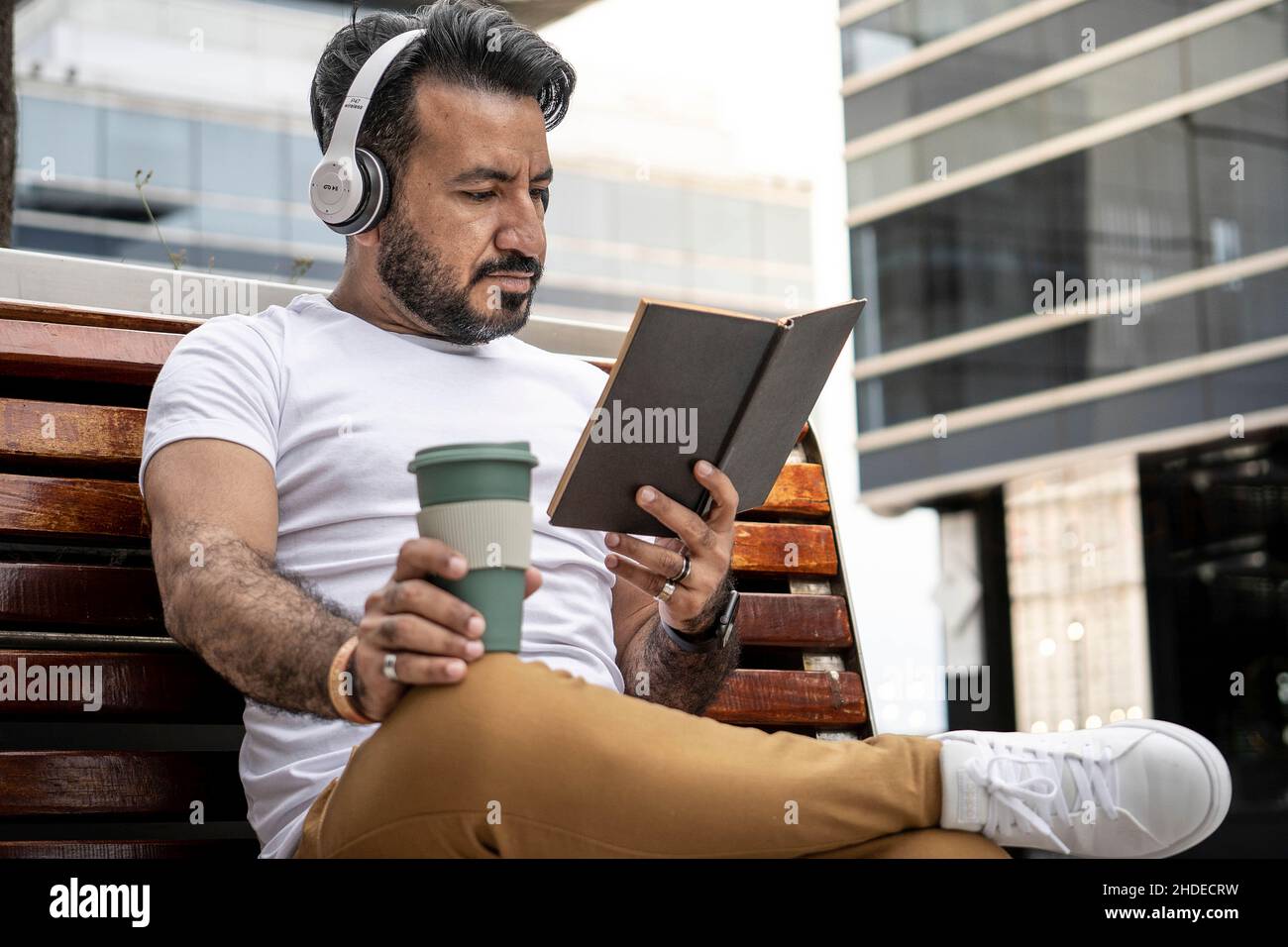 lateinischer Mann mit Bart sitzt auf einer Bank und liest ein Buch mit einer Tasse Kaffee in einer städtischen Umgebung. Stockfoto