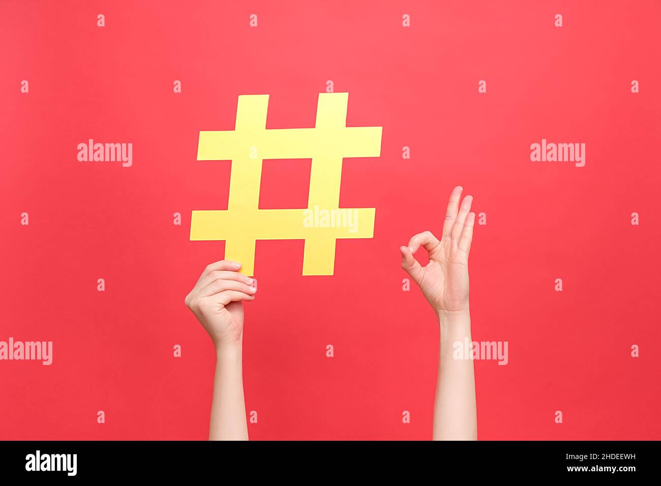 Weibliche Hände halten großen gelben Hashtag und zeigen den Finger darauf, okay Zeichen, isoliert über roten Studio Hintergrundwand. Social-Media-Markt Stockfoto