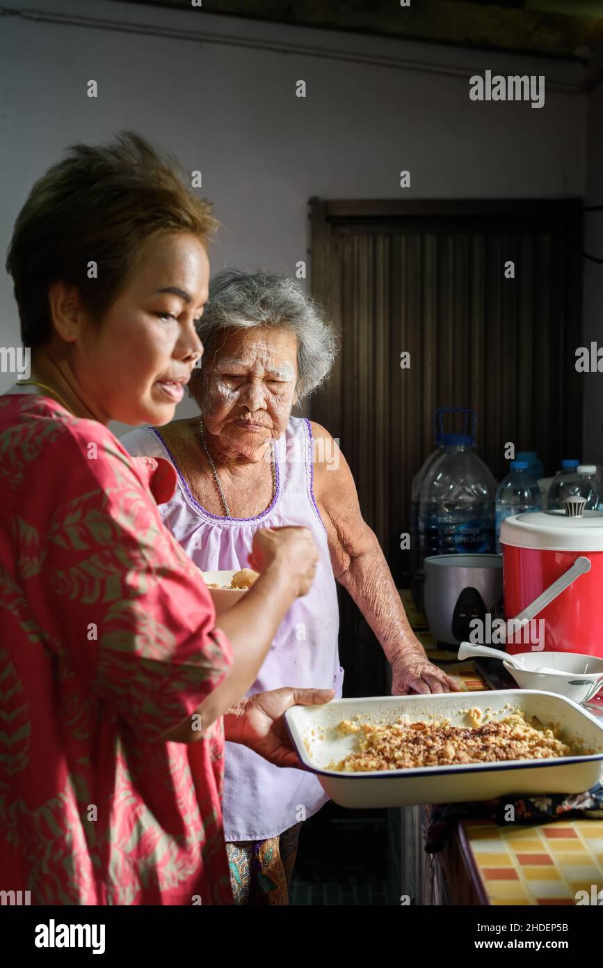 Mama und Oma stehen und halten Tablett mit frisch gebackenen Apfelkuchen in der Küche. Zwei Generationen thailändischer Mutter und Tochter, die Dessert kochen Stockfoto